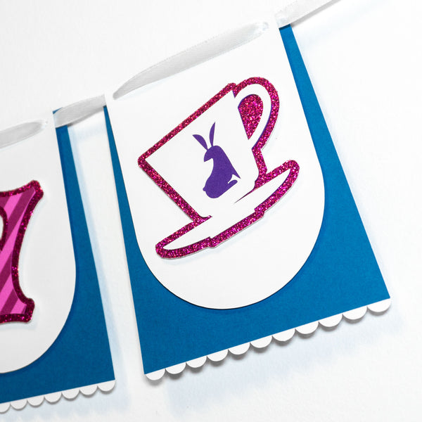Close-up of a handmade Alice in Wonderland “HAPPY BIRTHDAY” banner showing one full pennant & part of another. The individual paper pennants resembles a blue skirt & white apron, just like Alice's.  The partially visibile skirt-like pennant has the letter “Y” (of "happy") in a whimsical font & is striped pink like the Cheshire Cat. The main pennant pictured follows the "Y" and features a teacup with a rabbit motif on a glittery background. The pennants are strung together on a white satin ribbon