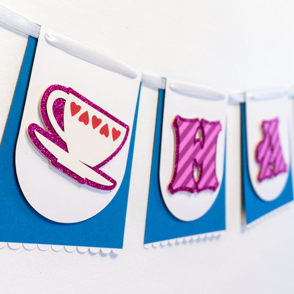Close-up of a handmade Alice in Wonderland “HAPPY BIRTHDAY” banner showing only the first three pennants. The individual paper pennants resembles a blue skirt & white apron, just like Alice's. The first skirt-like pennant pictured features a teacup with a heart motif on a glittery background. The next two pennants have the letters “H” and “A” in a whimsical font & are striped pink like the Cheshire Cat. They are strung together on a white satin ribbon.