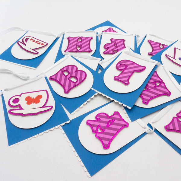 A handmade Alice in Wonderland “HAPPY BIRTHDAY” banner piled up on a white surface. Individual paper pennants can be seen and resemble blue skirts & white aprons, just like Alice's.  The skirt-like pennants feature either a letter or a teacup, both on a glittery background. The letters spell "HAPPY BIRTHDAY" and are in a pink-striped whimsical font like the Cheshire Cat. One of the teacup has a butterfly, the other has a heart motif.