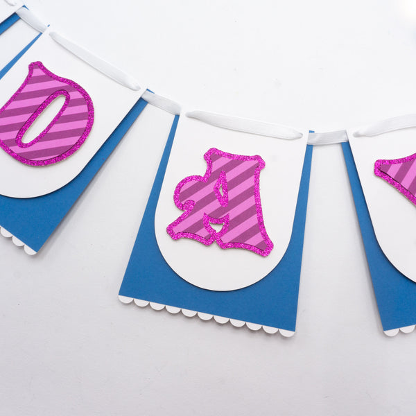 Close-up of a handmade Alice in Wonderland “HAPPY BIRTHDAY” banner showing parts of three individual pennants. Each paper pennants resembles a blue skirt & white apron, just like Alice's. The skirt-like pennants shown feature the letters D-A-Y (of "birthday") in a pink-striped whimsical font reminiscent of the Cheshire Cat. The letters are on a glittery background. The pennants are strung together on a white satin ribbon.