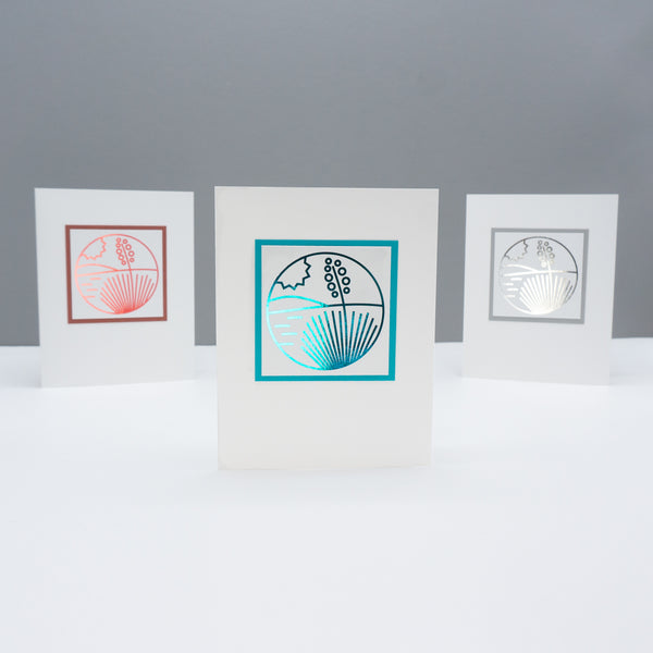 Three note cards all with the same modern monoline style drawing of a yucca in the desert with a hill in the background and the sun in the sky. The line drawing on each card is foiled in a different metallic color: terra cotta, turquoise, and silver.