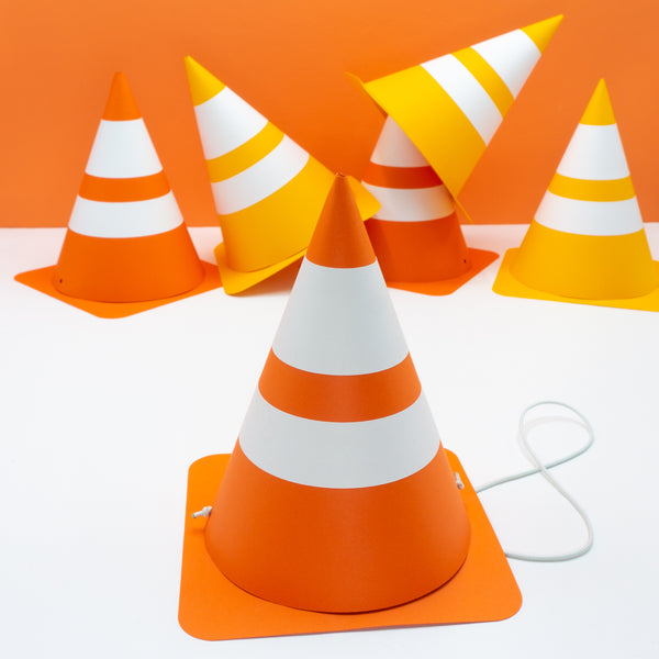 Handmade cardstock party hats that look like traffic cones. They are in two shades of orange. The hat in the front shows the fabric covered elastic strap that holds the hat on.
