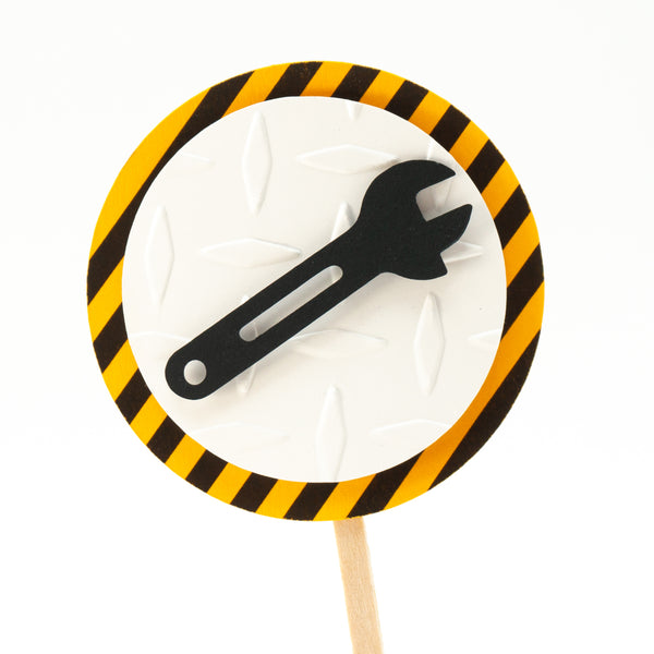 A cupcake toppers with a black wrench on top of a white circle of embossed diamond plate cardstock, which is placed on top of a circle of warning stripe paper.