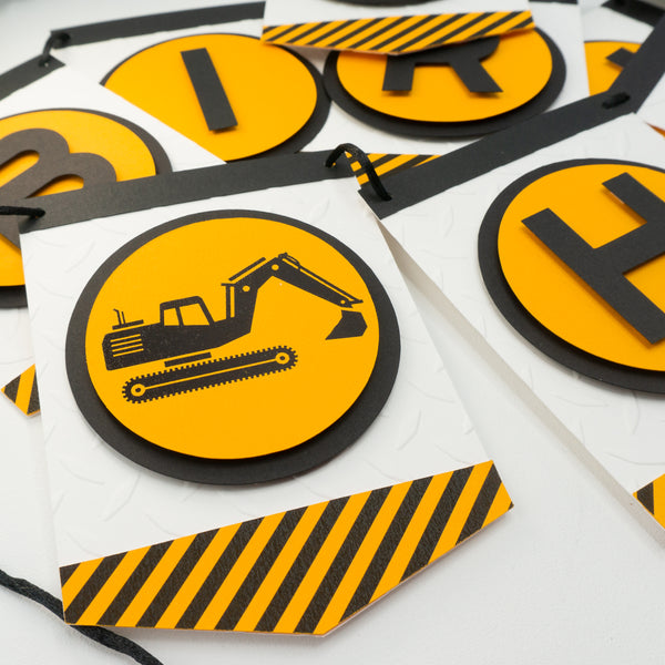 A close-up of a construction zone themed happy birthday banner. This photo shows off the silhouette of a backhoe which appears at the front of the “Happy” section of the banner. Each letter is in an orange circle and on a separate pennant. Strung on a black cord.