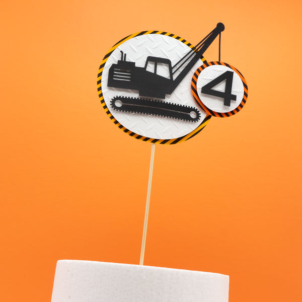 A construction zone cake topper featuring a silhouette of crawler crane which is hoisting a number representing the age of the birthday person. The crane and the number are on placed on a circle of diamond plate embossed white cardstock, which is on top of a circle of warning stipe cardstock.