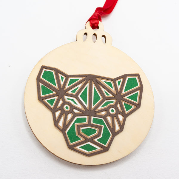 A close-up of a Christmas ornament with the face of a ferret. The design is in a modern geometric style featuring brown lines and filled in with green. It is cut from paper and attached to a wooden ornament-shaped disc. Hung on red ribbon.