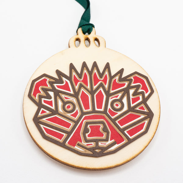 A close-up of a Christmas ornament with the face of a hedgehog. The design is in a modern geometric style featuring brown lines and filled in with red. It is cut from paper and attached to a wooden ornament-shaped disc. Hung on green ribbon.