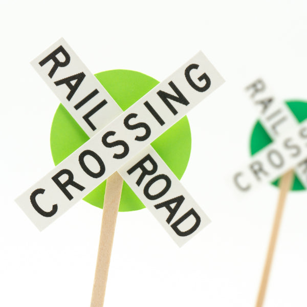 A closeup of one cupcake topper with another out of focus in the background. The toppers consist of the crossbuck style railroad crossing sign with a smaller circle of green behind them as an accent color.