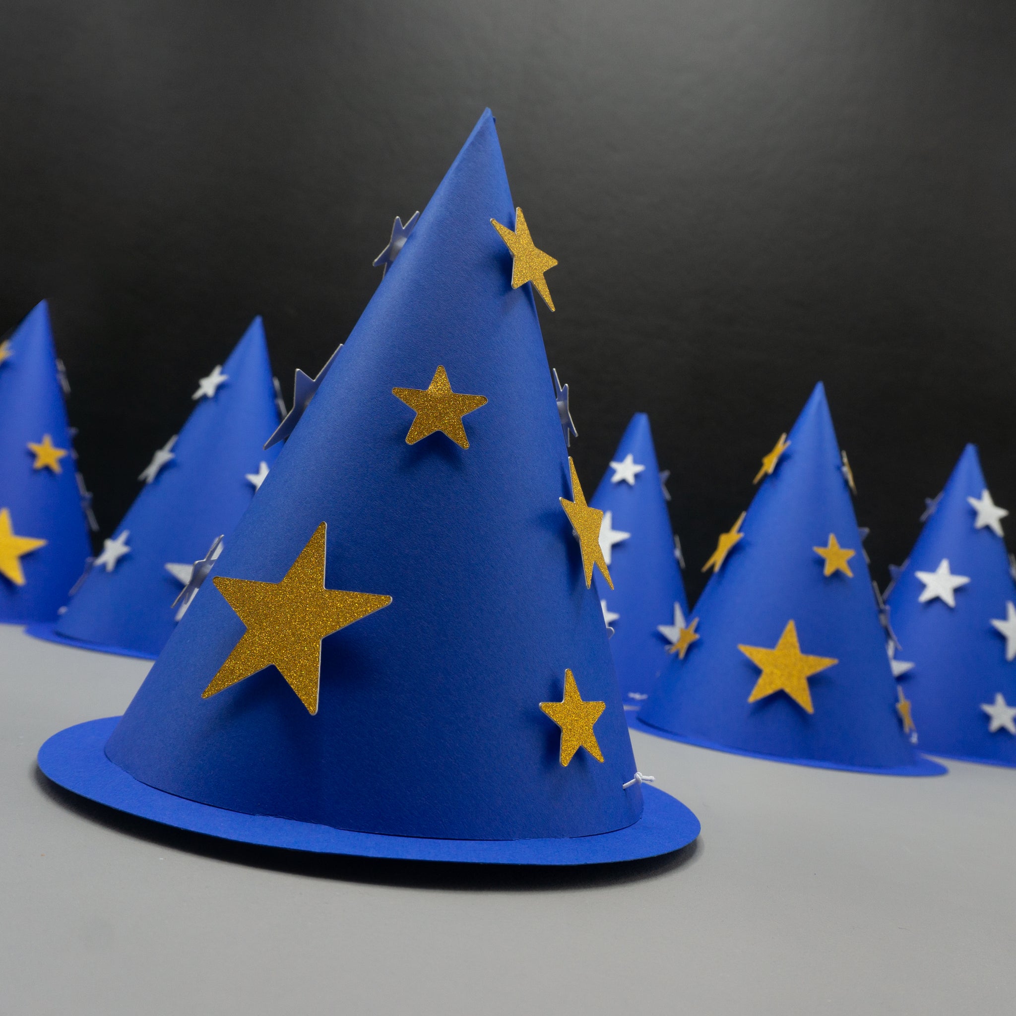 foreground hat is dark blue with shimmering gold stars, while the five hats in the background are slightly out of focus—two with gold stars and three with silver. These cone hats combine the look of traditional party hats with magical wizard costume accessories.