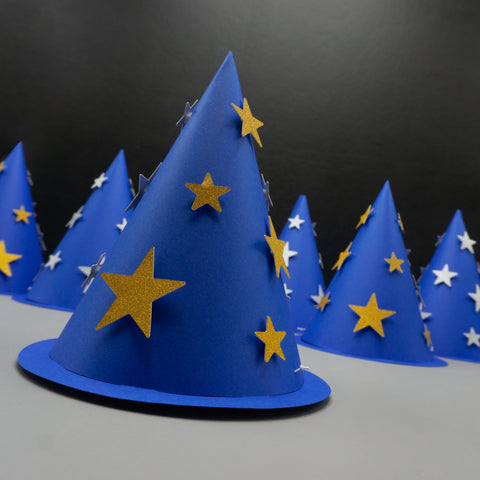 foreground hat is dark blue with shimmering gold stars, while the five hats in the background are slightly out of focus—two with gold stars and three with silver. These cone hats combine the look of traditional party hats with magical wizard costume accessories.