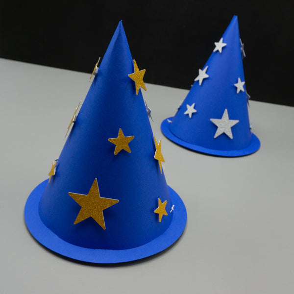 Two blue cone-shaped wizard party hats with thin brims rest on a gray surface against a black background. The hat in the foreground is decorated with scattered gold glitter stars of various sizes, while the hat in the background features silver glitter stars. Both resemble classic wizard costume accessories styled like party hats.