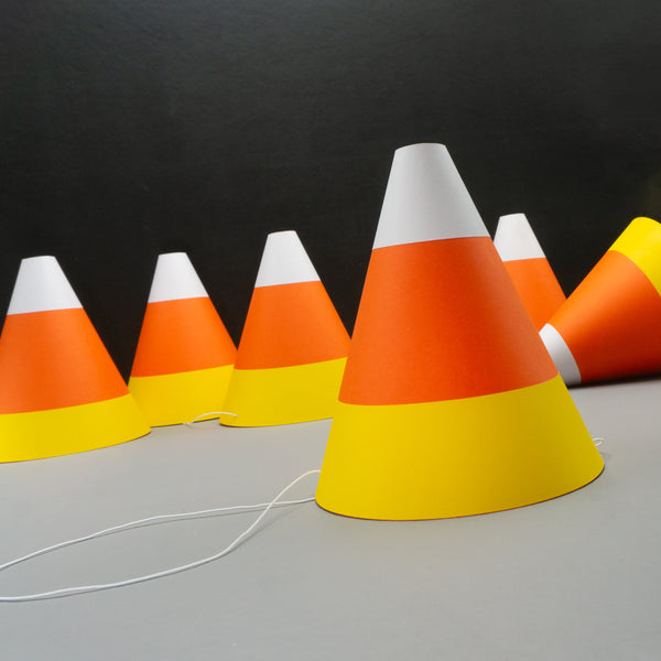 Six cone-shaped paper party hats designed to look like candy corn, the classic Halloween treat, with white, orange, and yellow stripes, arranged on a gray surface against a black background.