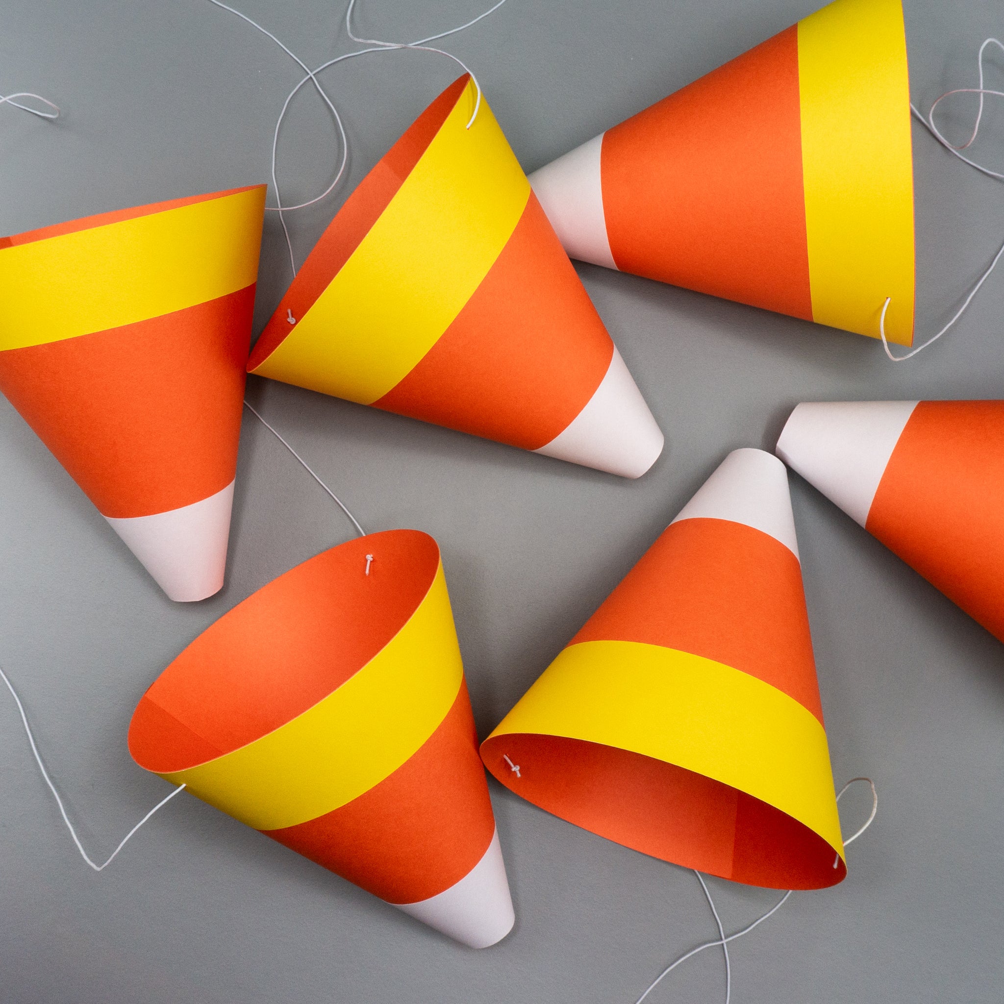 Six cone-shaped paper party hats designed to look like candy corn, the classic Halloween treat, with white, orange, and yellow stripes. The hats lie on their sides on a gray surface, scattered like a handful of candy. White elastic bands, used to secure the costume accessories, are visible.