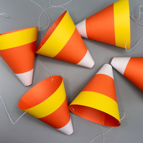 Six cone-shaped paper party hats designed to look like candy corn, the classic Halloween treat, with white, orange, and yellow stripes. The hats lie on their sides on a gray surface, scattered like a handful of candy. White elastic bands, used to secure the costume accessories, are visible.