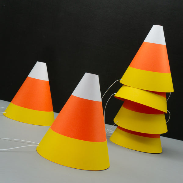 Six cone-shaped paper party hats designed to look like candy corn, the classic Halloween treat, with white, orange, and yellow stripes. One hat is in focus in the foreground, while five are stacked in the background against a black surface. White elastic bands, used to secure the costume accessories, are visible.