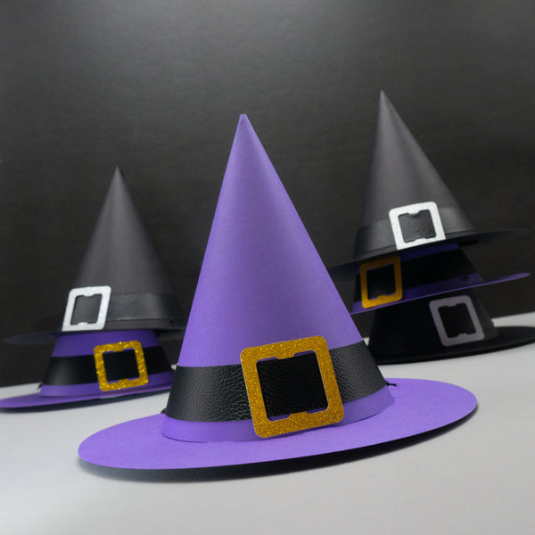 Six standard cone-shaped paper party hat, resembling witch hats. Complete with wide brims and buckled hat bands. The hat in the foreground is purple, with a black pleather band, and a gold glittery buckle. Five more hats are stacked in the background. Two of them are purple. The other three are all black with silver buckles.