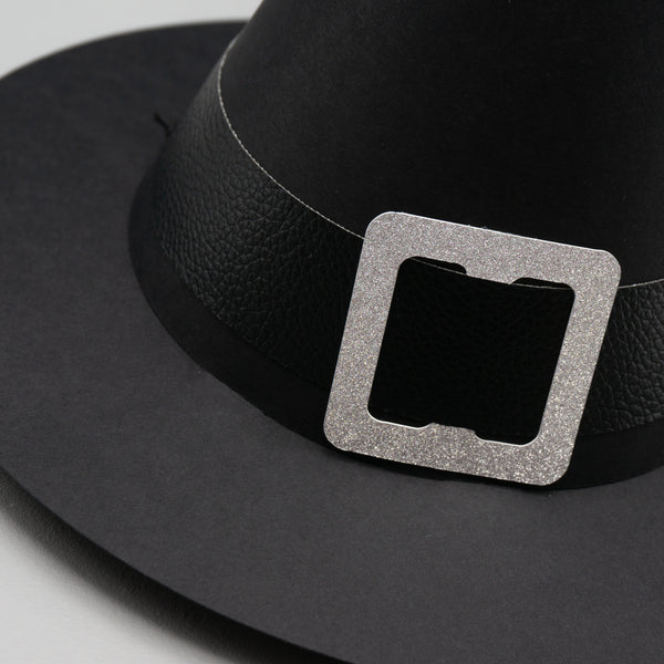 A close-up of standard cone-shaped paper party hat, resembling a witch hat. Complete with a wide brim and a buckled hat band. The hat is black with a black pleather band and a silver glittery buckle. The leather texture is visible on the hat band.