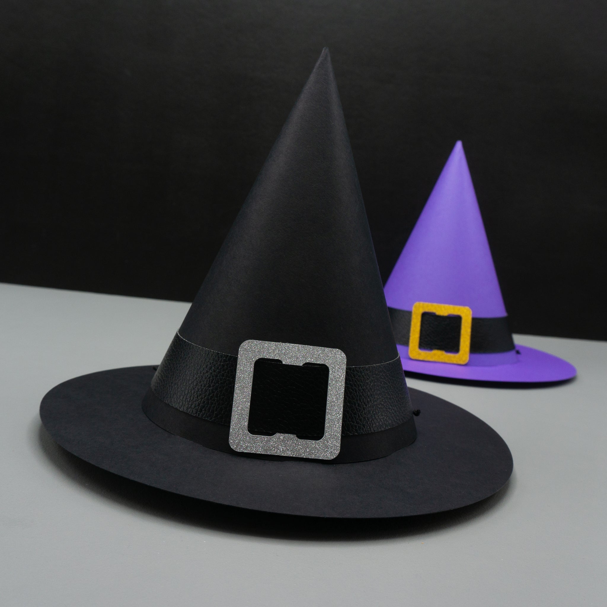 Two standard cone-shaped paper party hat, resembling witch hats. Complete with wide brims and buckled hat bands. The hat in the foreground is black, with a black pleather band, and a silver glittery buckle. The hat in the background is purple, with a black pleather band, and a gold glittery buckle. 