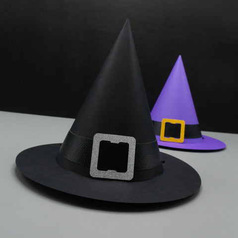 Two standard cone-shaped paper party hat, resembling witch hats. Complete with wide brims and buckled hat bands. The hat in the foreground is black, with a black pleather band, and a silver glittery buckle. The hat in the background is purple, with a black pleather band, and a gold glittery buckle. 