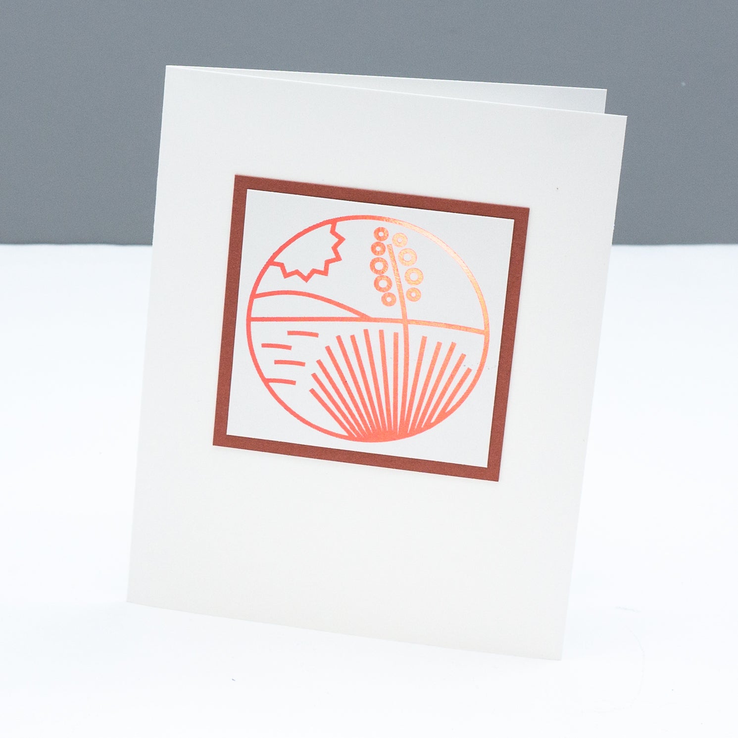 Note card with a modern monoline style drawing of a yucca in the desert with a hill in the background and the sun in the sky. the line drawing is foiled in a metallic terra cotta color. 