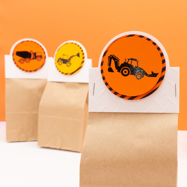 Small paper bag party favor bags with construction zone themed header cards attached. Header cards feature shiny black construction equipment on bright orange backgrounds surrounded by a border of warning stripes.
