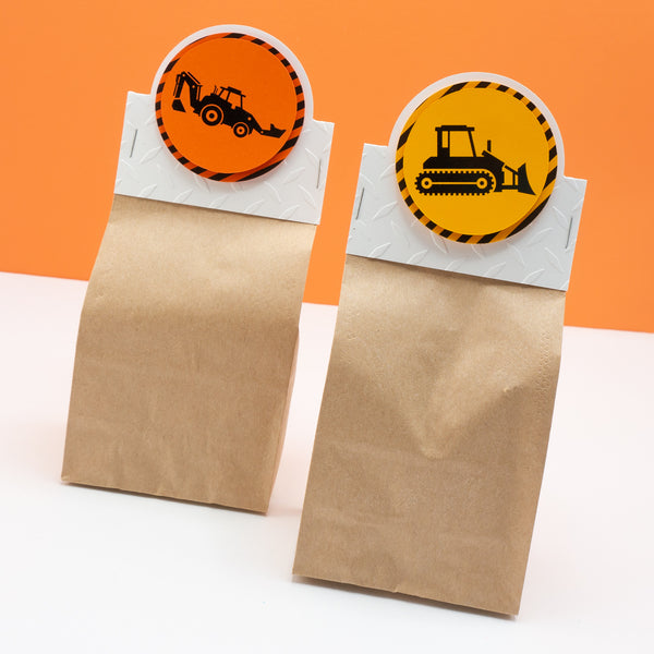 Small paper bag party favor bags with construction zone themed header cards attached. Header cards feature shiny black construction equipment on bright orange backgrounds surrounded by a border of warning stripes.