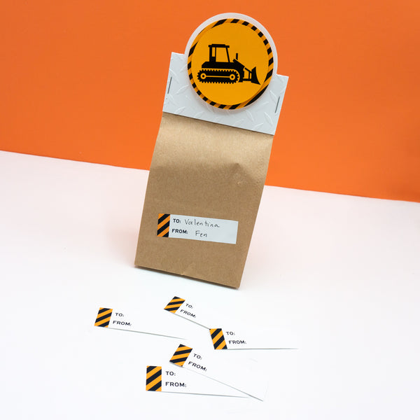 One small paper bag party favor bag with construction zone themed header card attached. Header card features a shiny black bulldozer on bright golden backgrounds surrounded by a border of warning stripes. A gift tag with an edge of caution tape is affixed the the bag.