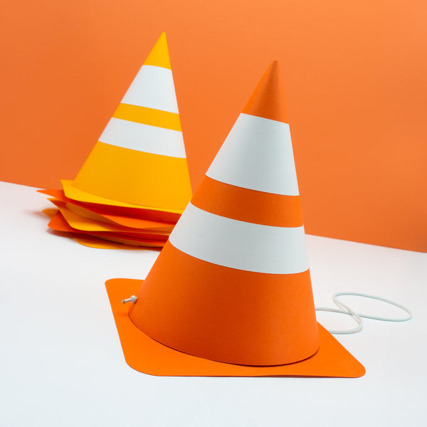 Handmade cardstock party hats that look like traffic cones. They are in two shades of orange. The hat in the front shows the fabric covered elastic strap that holds the hat on.