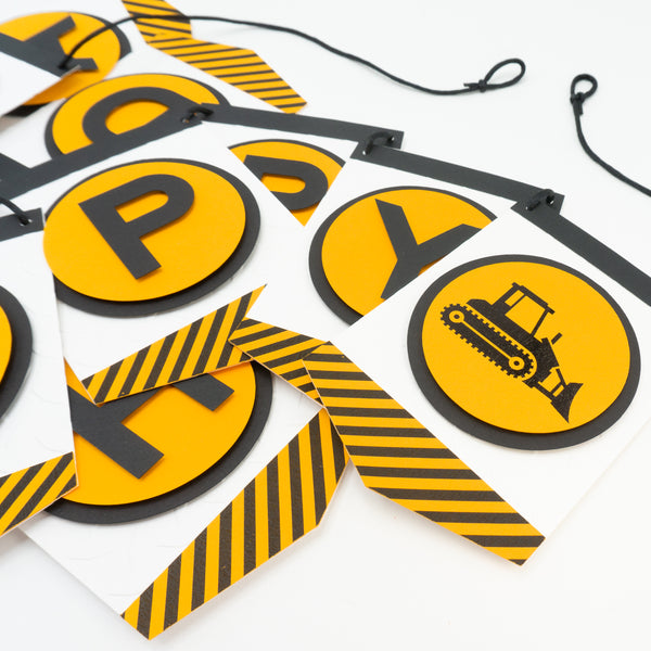 A close-up of a construction zone themed happy birthday banner. This photo shows off the silhouette of a bulldozer which appears at the end of the “Happy” section of the banner. Each letter is in an orange circle and on a separate pennant. Strung on a black cord.