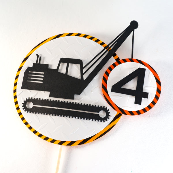 A construction zone cake topper featuring a silhouette of crawler crane which is hoisting a number representing the age of the birthday person. The crane and the number are on placed on a circle of diamond plate embossed white cardstock, which is on top of a circle of warning stipe cardstock.