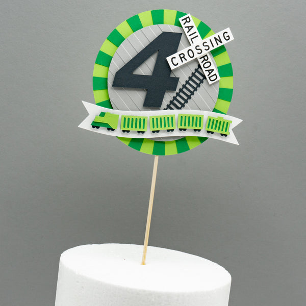 A circular cake topper with a burst of green surrounding it. A short freight train in a small banner is affixed to the bottom of the topper. A large number four in the center is accented with a railroad crossing sign and a section of train tracks.