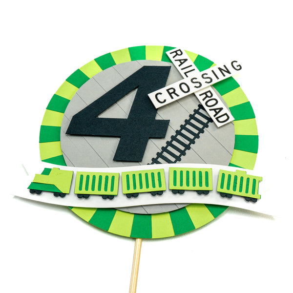 A circular cake topper with a burst of green surrounding it. A short freight train in a small banner is affixed to the bottom of the topper. A large number four in the center is accented with a railroad crossing sign and a section of train tracks. This photos is a closer view to show off the layering of the paper.