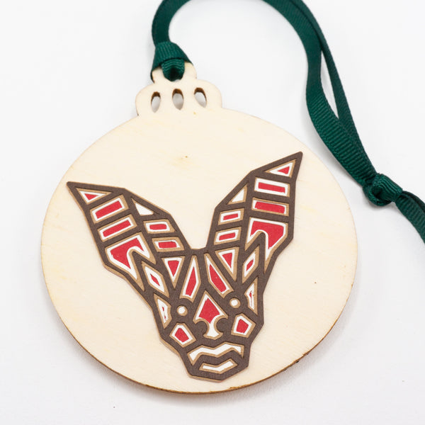 A close-up of a Christmas ornament with the face of a large-eared bat. The design is in a modern geometric style featuring brown lines and filled in with red. It is cut from paper and attached to a wooden ornament-shaped disc. Hung on green ribbon.