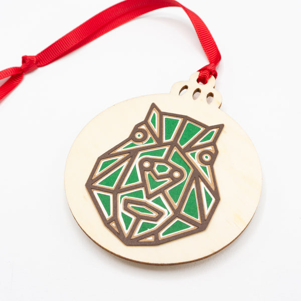 A close-up of a Christmas ornament with the face of a capybara. The design is in a modern geometric style featuring brown lines and filled in with green. It is cut from paper and attached to a wooden ornament-shaped disc. Hung on red ribbon.