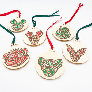 Six Christmas ornaments, each with a different animal face. The faces are made in a modern geometric style featuring brown lines and filled in with red or green. the design is cut from paper and is attached to a wooden ornament-shaped disc.