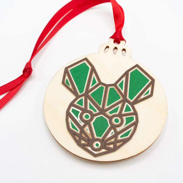 A close-up of a Christmas ornament with the face of a mouse. The design is in a modern geometric style featuring brown lines and filled in with green. It is cut from paper and attached to a wooden ornament-shaped disc. Hung on red ribbon.