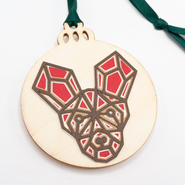 A close-up of a Christmas ornament with the face of a painted dog. The design is in a modern geometric style featuring brown lines and filled in with red. It is cut from paper and attached to a wooden ornament-shaped disc. Hung on green ribbon.