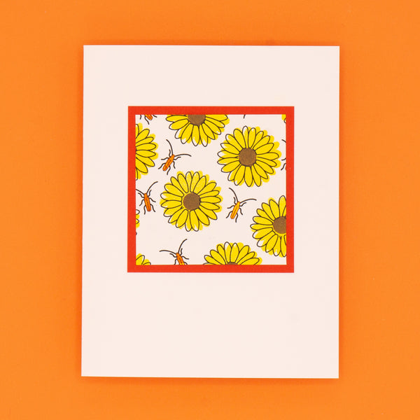 A straight-on photograph of a closed notecard. The card is adorned with a square of a bold, stamped, yellow sunflowers pattern. The flowers are interspersed with metallic orange beetles. The patterned square is surrounded by a thin orange border.