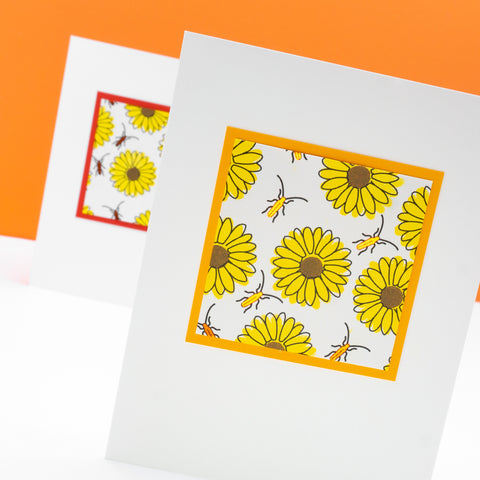 A photograph of a notecard in the foreground, with another notecard behind it and slightly out of focus. On each card an orange border surrounds a patterned square of stamped yellow sunflowers interspersed with orange metallic beetles.