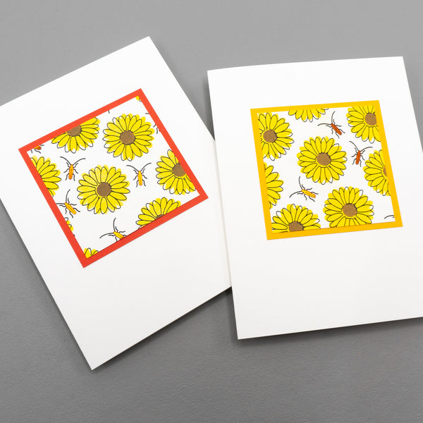 Two notecards lying on a surface, photographed from above. Both cards are decorated with a square of bold stamped yellow sunflowers pattern. The flowers are interspersed with metallic orange beetles. The patterned square is surrounded by a thin orange border.