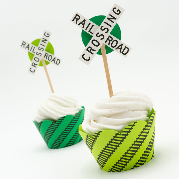 Two cupcake toppers stuck in cupcakes. The toppers consist of the crossbuck style railroad crossing sign with a smaller circle of green behind them as an accent color.