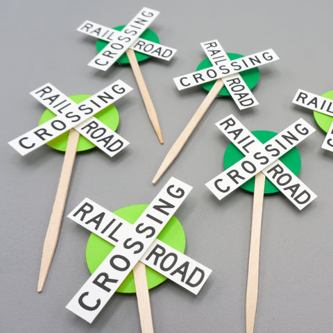 Several cupcake toppers lying flat. They consist of the crossbuck style railroad crossing sign with a smaller circle of green behind them as an accent color.